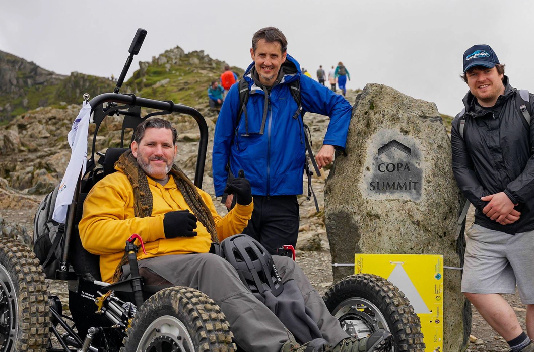 Nick Wilson in RockClimber beside the Copa Summit sign on Mount Snowdon. Nick is smiling, wearing a yellow hoodie, grey trousers, and black gloves. Nick Wilson in RockClimber beside the Copa Summit sign on Mount Snowdon. Nick is smiling, wearing a yellow hoodie, grey trousers, and black gloves.