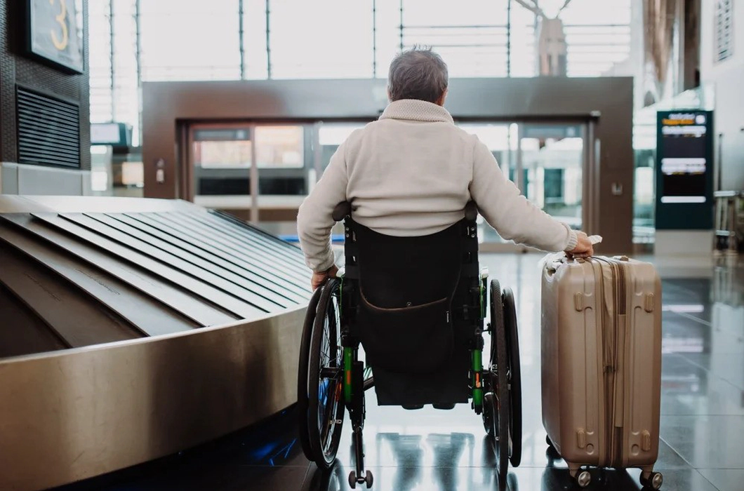 A man wearing a white jumper sitting in a wheelchair with his back to the camera, holding a suitcase on wheels in his right hand, in an airport. A man wearing a white jumper sitting in a wheelchair with his back to the camera, holding a suitcase on wheels in his right hand, in an airport.