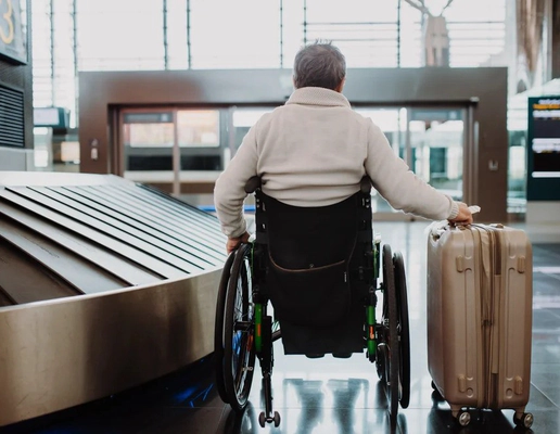 A man wearing a white jumper sitting in a wheelchair with his back to the camera, holding a suitcase on wheels in his right hand, in an airport. A man wearing a white jumper sitting in a wheelchair with his back to the camera, holding a suitcase on wheels in his right hand, in an airport.