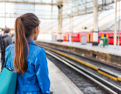 Photo of a train platform. The platform and people on the platform are blurred except for a woman in the foreground who stands with her back to the camera with long brown hair in a ponytail and a blue denim jacket. A red train is waiting at the platform opposite. Photo of a train platform. The platform and people on the platform are blurred except for a woman in the foreground who stands with her back to the camera with long brown hair in a ponytail and a blue denim jacket. A red train is waiting at the platform opposite.