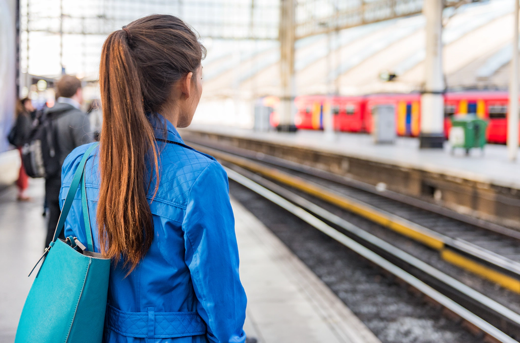 Photo of a train platform. The platform and people on the platform are blurred except for a woman in the foreground who stands with her back to the camera with long brown hair in a ponytail and a blue denim jacket. A red train is waiting at the platform opposite. Photo of a train platform. The platform and people on the platform are blurred except for a woman in the foreground who stands with her back to the camera with long brown hair in a ponytail and a blue denim jacket. A red train is waiting at the platform opposite.